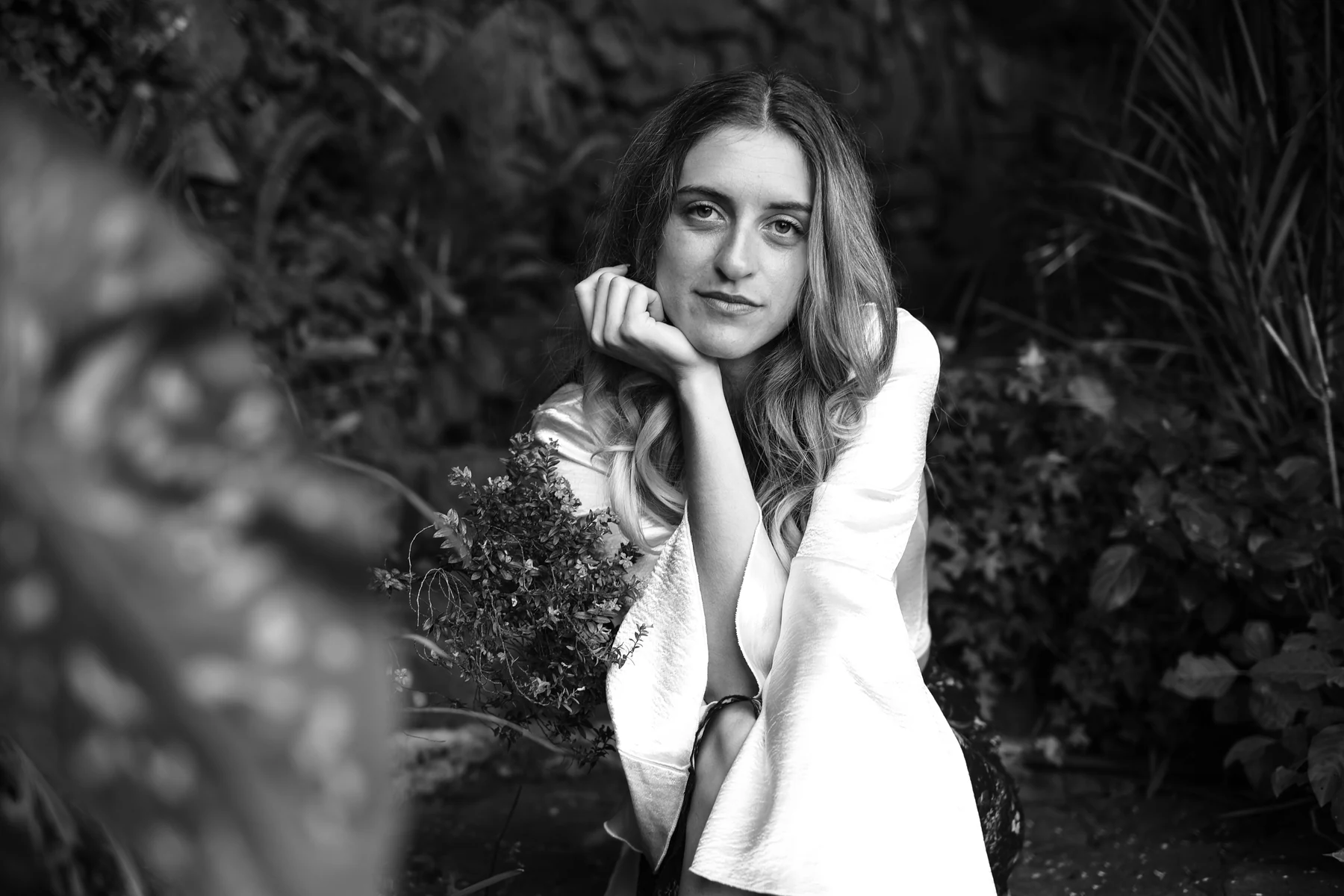 Black-and-white portrait of Marissa looking toward the camera, her chin resting on her hand in a garden.