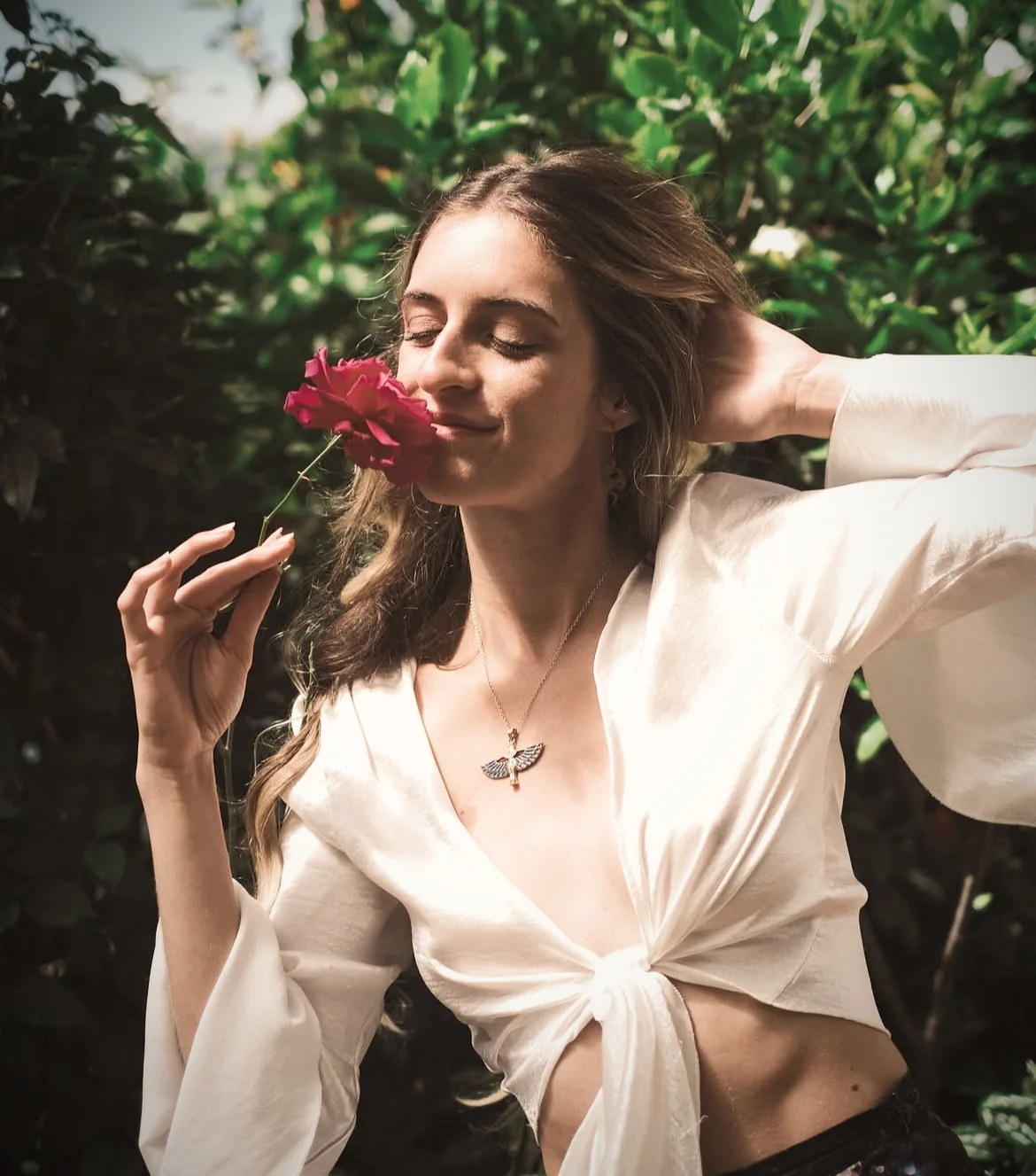 Marissa in a cream blouse, smelling a red rose in a leafy garden.