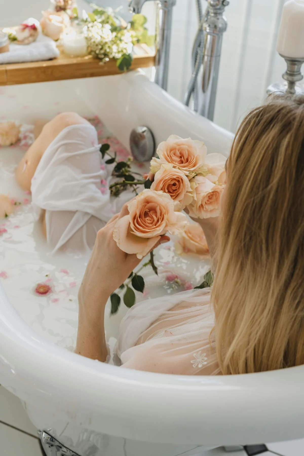 A woman holding peach roses while resting in a milky bath scattered with petals.