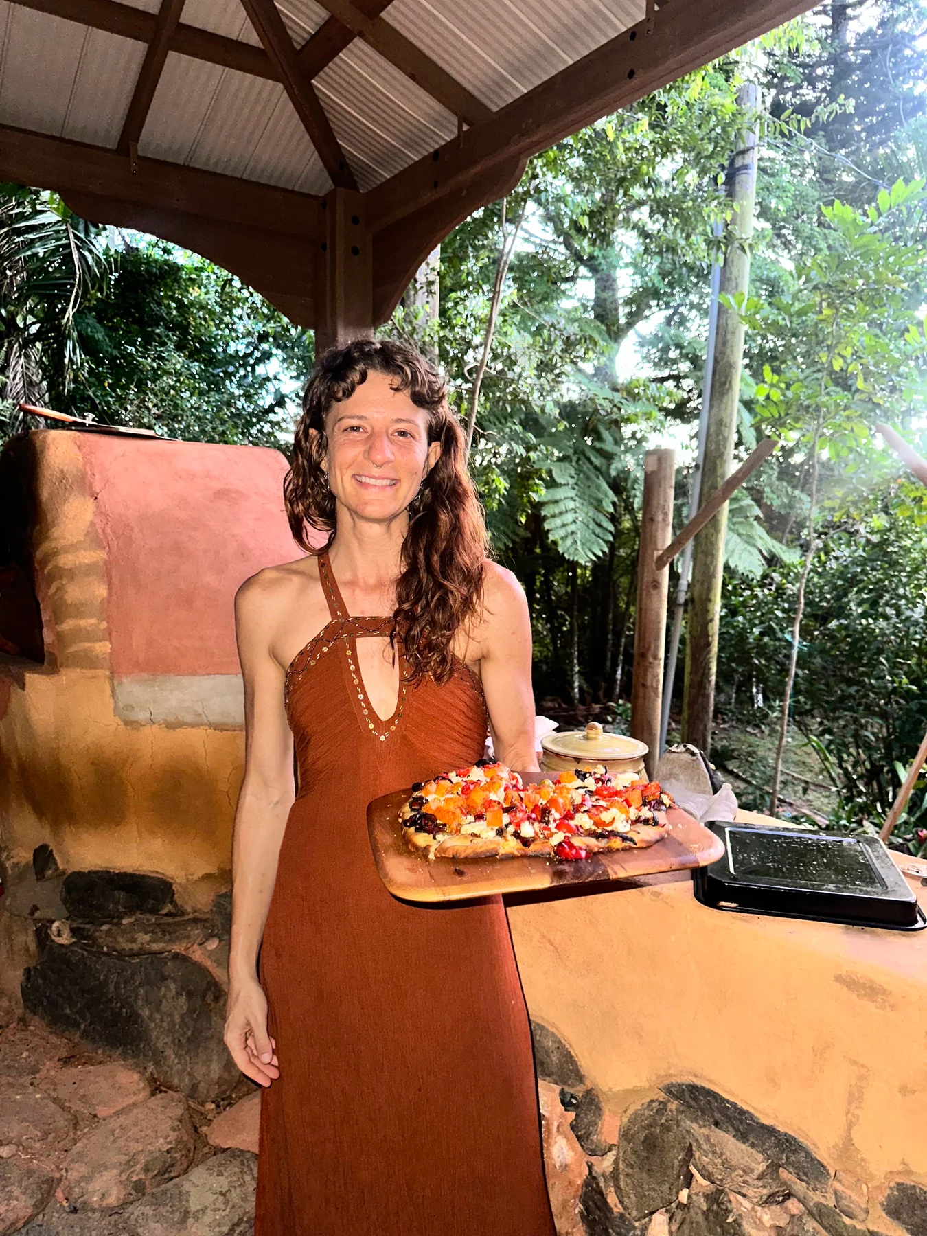 Charity Luce Colón smiling beside an earthen oven, holding a wooden board of freshly prepared food.