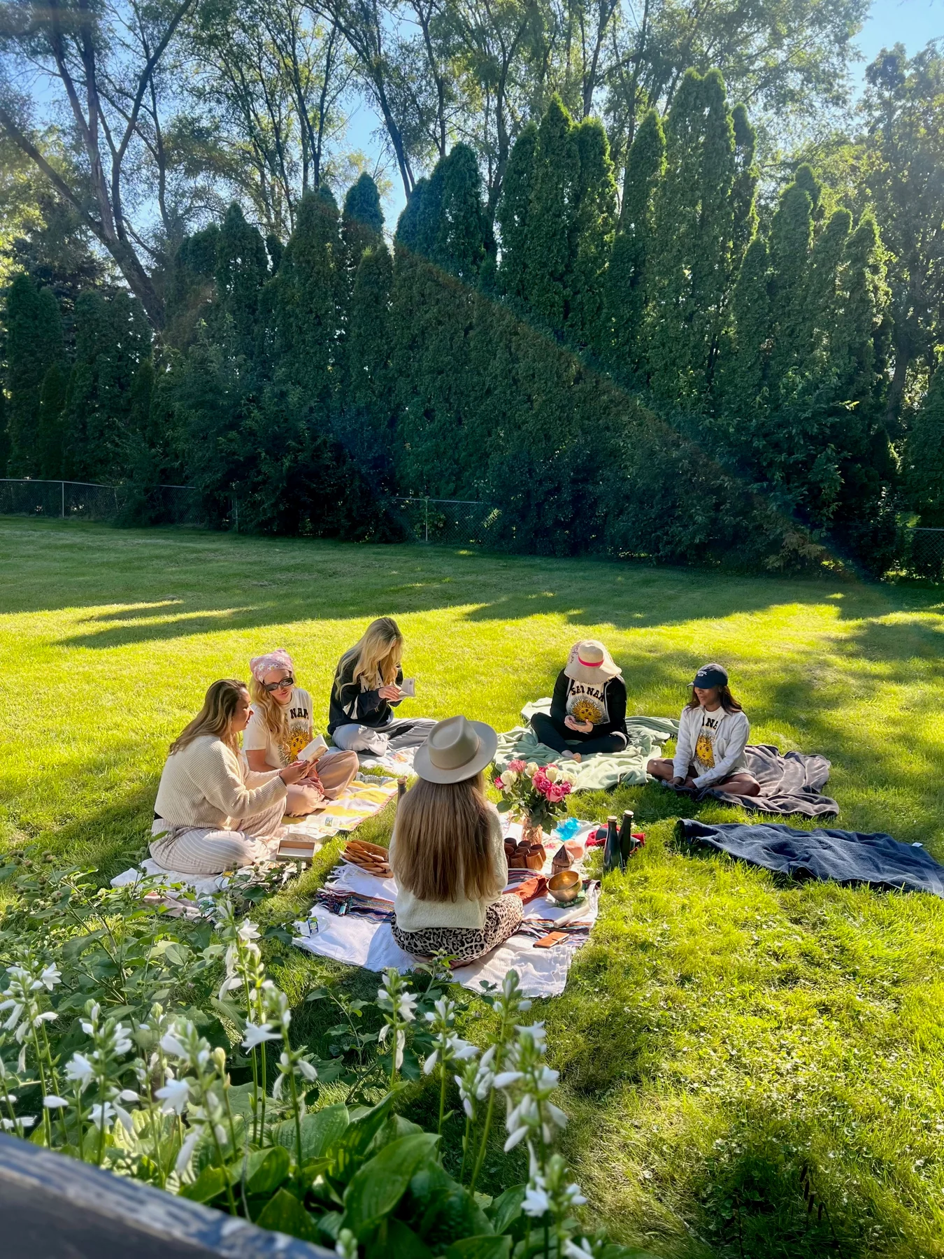 Women gathered in a circle on blankets in a sunlit garden, with flowers and ritual objects between them.